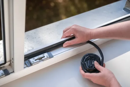 د یوه کس انځور چې د کړکۍ پر څنډه ربړي ربړي پټۍ (weatherstripping) نصبوي.