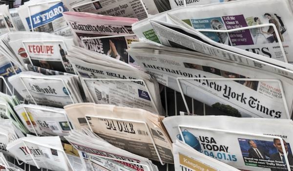 A display rack filled with various newspapers, including The New York Times, USA Today, and other international and local publications. The newspapers are arranged in an overlapping manner, showcasing different headlines and front-page stories.