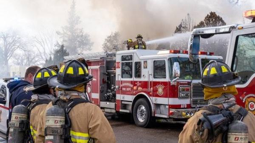 Firefighters in full gear are working at the scene of a fire. Several firefighters are facing a fire truck, with one standing on top, directing a hose at the burning building. Smoke is billowing in the background, and additional emergency vehicles are present.