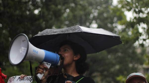 A woman is holding a megaphone and speaking into it while standing under an umbrella in the rain. She appears to be at an outdoor event or protest, with blurred greenery in the background.