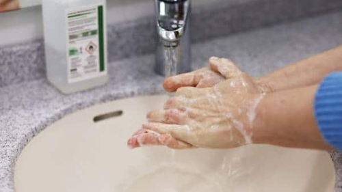 Hands covered in soap lather are being washed under running water in a sink. A bottle of liquid soap is visible on the counter next to the sink.