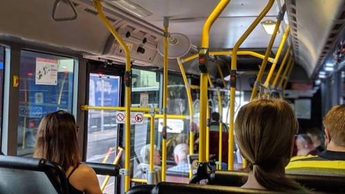 The interior of a city bus with passengers seated and some standing, holding onto yellow handrails. The view is towards the front of the bus, showing the driver and the bus's entrance. The bus is in motion, passing through an urban area.