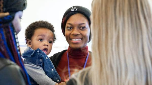 A smiling woman wearing a black hat is holding a baby and standing in a group with other women. The baby is looking curiously at one of the women in the group. The setting appears to be casual and friendly.