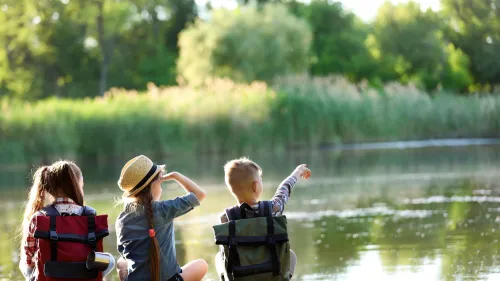 Three children sitting on a dock next to a body of water. One child is pointing across the water.