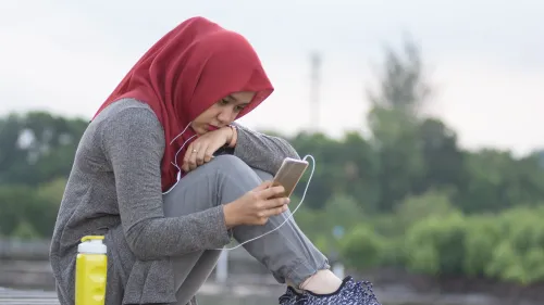 Photo of a teenager sitting with headphones in her ears and looking at her cell phone.
