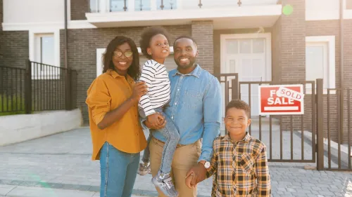 A family of four is standing in front of a home that has a sold sign on the front gate.