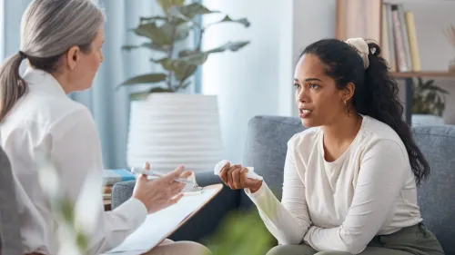 A person who appears to be upset is holding a tissue and sitting on a couch while speaking with a counselor who is taking notes on a clipboard