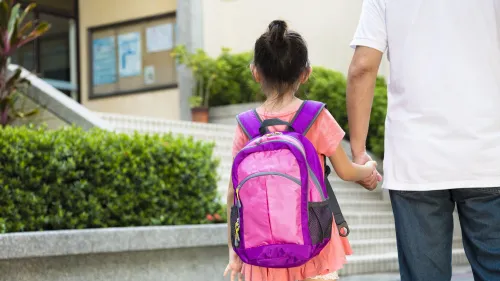 Un adulto lleva de la mano a su hija al entrar al colegio. La niña lleva una mochila. 