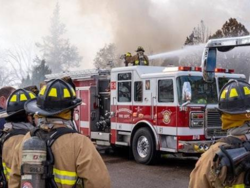 Firefighters in full gear are working at the scene of a fire. Several firefighters are facing a fire truck, with one standing on top, directing a hose at the burning building. Smoke is billowing in the background, and additional emergency vehicles are present.