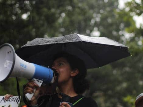 A woman is holding a megaphone and speaking into it while standing under an umbrella in the rain. She appears to be at an outdoor event or protest, with blurred greenery in the background.