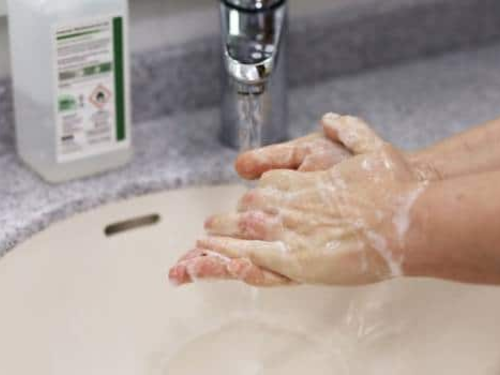 Hands covered in soap lather are being washed under running water in a sink. A bottle of liquid soap is visible on the counter next to the sink.