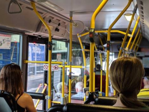 The interior of a city bus with passengers seated and some standing, holding onto yellow handrails. The view is towards the front of the bus, showing the driver and the bus's entrance. The bus is in motion, passing through an urban area.