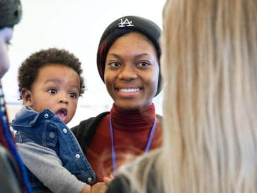 A smiling woman wearing a black hat is holding a baby and standing in a group with other women. The baby is looking curiously at one of the women in the group. The setting appears to be casual and friendly.