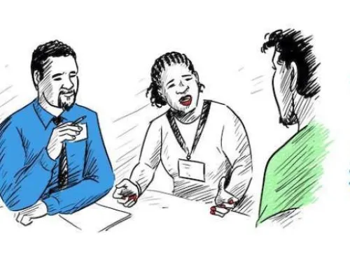 Three people are engaged in a discussion. A man in a blue shirt is holding a pen, a woman with braided hair and a name tag is speaking animatedly, and a man with curly hair in a green shirt is listening. The setting appears to be a meeting or consultation.