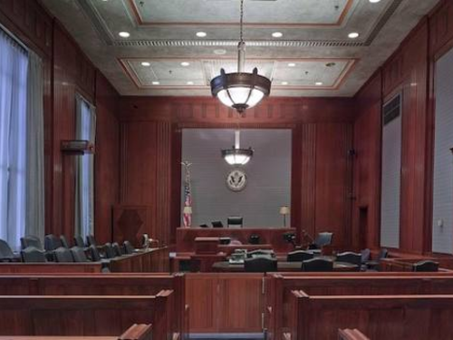 An empty courtroom featuring wooden furnishings, a judge's bench, jury seats, and a witness stand. The room is well-lit with chandeliers hanging from the ceiling, and an American flag is visible behind the judge's bench.