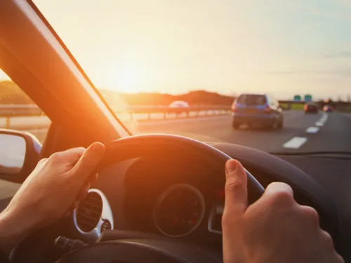 Photograph of driver with hands on steering wheel on a highway at sunset.