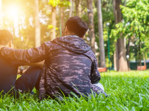 Photo of a man and a boy sitting in a grass field. The man is comforting the boy by putting his arm on the boy’s shoulder.