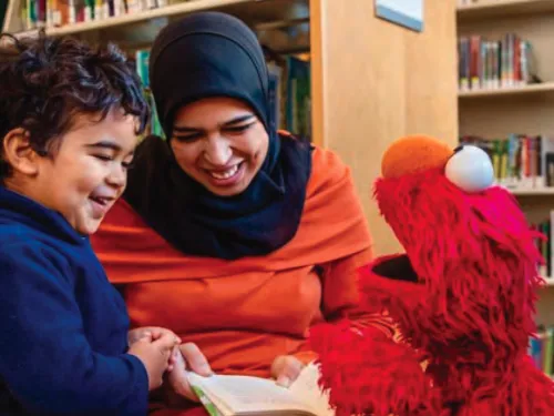 Photo of Afghan mother, child, and puppet from a television show smiling and reading a book in the library. Photo Credit: Sesame Workshop