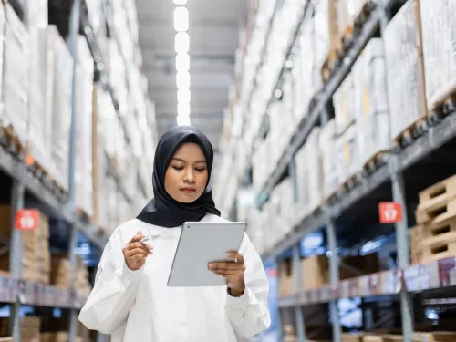 Photo of a woman working in the aisle of a large warehouse with a computer tablet.