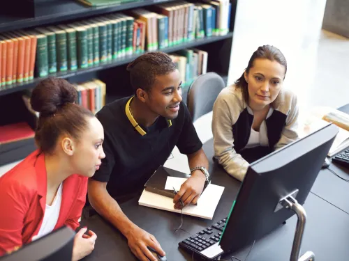 Three students look at a computer screen in a library with shelves of books in the background.