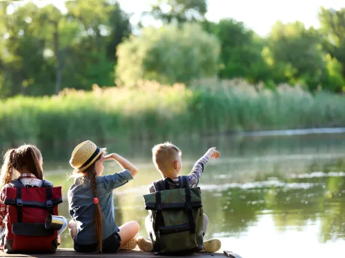 Three children sitting on a dock next to a body of water. One child is pointing across the water.