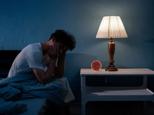 A man sitting on the edge of his bed in a dimly lit room with his hands holding his head. There is a lamp and alarm clock on the nightstand next to the bed.