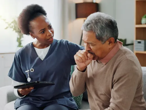 A healthcare provider offers support to a patient who is coughing into his hand while seated on a couch.