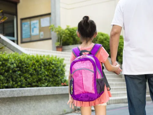 An adult is holding their child’s hand as they walk into the school building. The child is wearing a backpack.