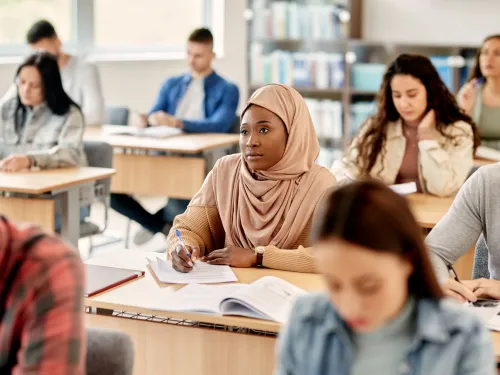 A female student wearing a hijab sits among other adult learners in a classroom.