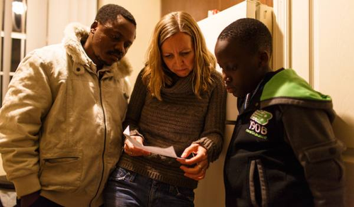 Three people are standing together in a room, closely looking at a piece of paper. A man in a white jacket, a woman with blonde hair in a gray sweater, and a boy in a black and green jacket are intently reading the document. The setting appears to be a casual indoor environment.
