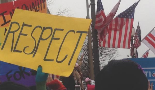 A crowd of people holding signs at a protest or rally. The foreground sign is yellow and reads 'RESPECT.' In the background, several American flags are visible.