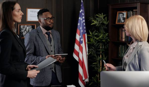 Three professionals are having a discussion in an office setting. A woman with long brown hair and a man with glasses and a beard, both holding tablets, are listening to a blonde woman who is speaking. An American flag stands in the background, along with bookshelves and framed photographs.
