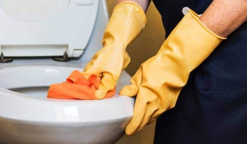 Person wearing yellow rubber gloves is cleaning a toilet with an orange cloth. The toilet lid is open, and the person is focused on scrubbing the rim of the toilet bowl.