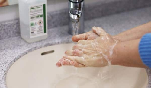 Hands covered in soap lather are being washed under running water in a sink. A bottle of liquid soap is visible on the counter next to the sink.