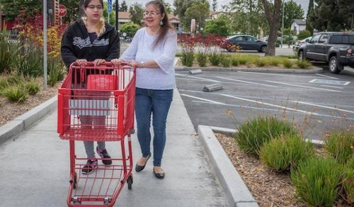 Two women are walking on a sidewalk in a parking lot, pushing a red shopping cart. One woman is wearing a black hoodie, and the other is wearing a white blouse. They appear to be having a conversation as they walk.