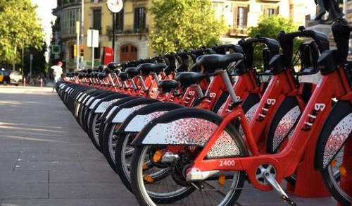 A row of red rental bicycles is lined up at a bike-sharing station in an urban area. The bikes are neatly arranged and ready for use. In the background, there are trees and buildings, suggesting the station is located in a city square or park.