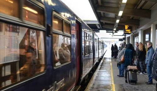 Passengers wait on a platform as a train arrives at a station. Some people are standing with luggage, while others are seated inside the train, visible through the windows. The platform is covered, and there is a digital sign displaying information about the train's arrival.
