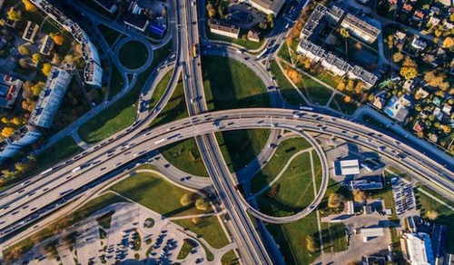 An aerial view of a complex highway interchange with multiple lanes, overpasses, and ramps. Cars are traveling in various directions. Surrounding the interchange are buildings, green spaces, and residential areas, creating a bustling urban landscape.