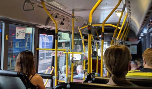 The interior of a city bus with passengers seated and some standing, holding onto yellow handrails. The view is towards the front of the bus, showing the driver and the bus's entrance. The bus is in motion, passing through an urban area.