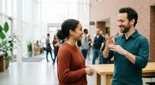 Two people smile and use sign language in a busy office lobby.