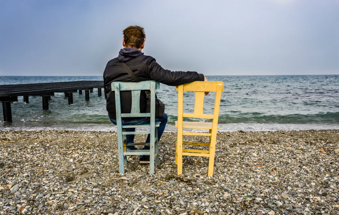 Photo of a man in a chair facing the sea beside an empty chair.