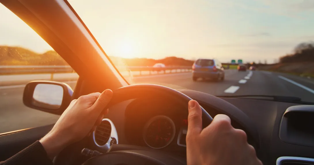 Photograph of driver with hands on steering wheel on a highway at sunset.