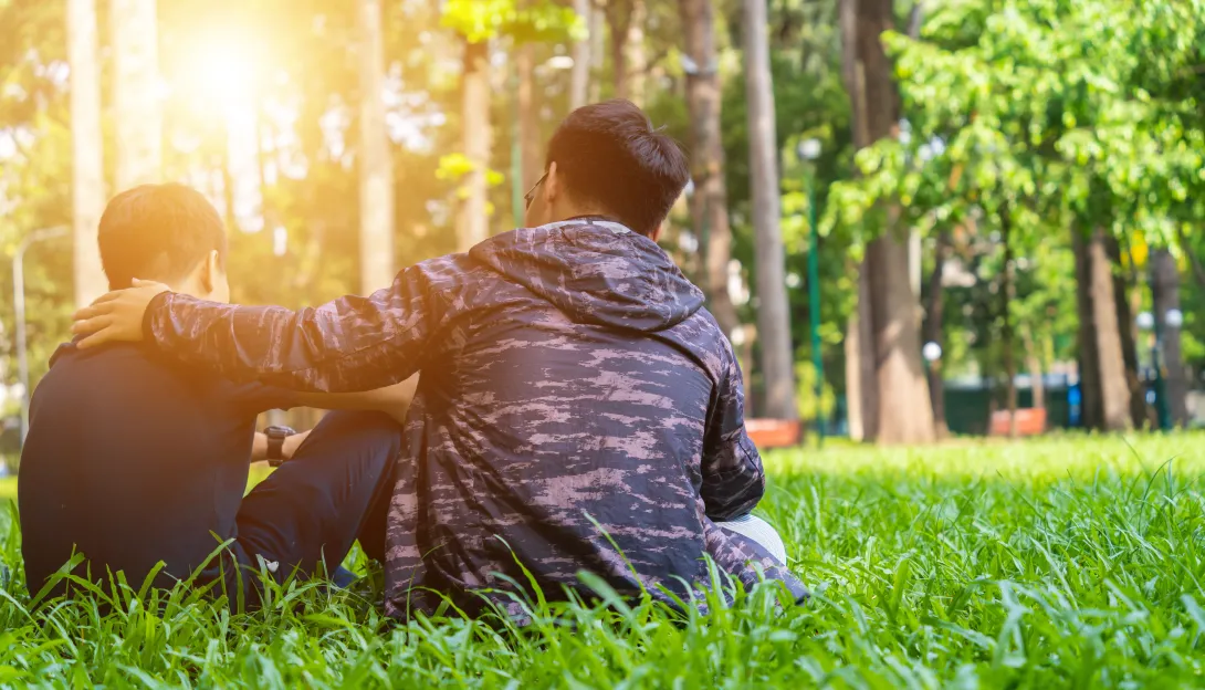 Photo of a man and a boy sitting in a grass field. The man is comforting the boy by putting his arm on the boy’s shoulder.