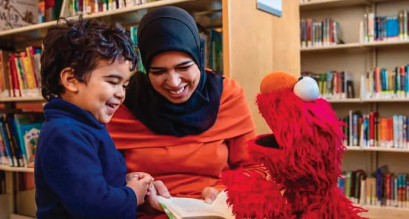 Photo of Afghan mother, child, and puppet from a television show smiling and reading a book in the library.  Photo Credit: Sesame Workshop