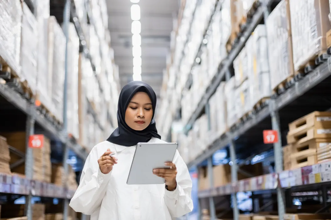 Photo of a woman working in the aisle of a large warehouse with a computer tablet.