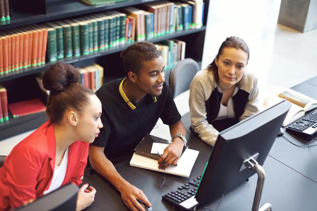 Three students look at a computer screen in a library with shelves of books in the background.
