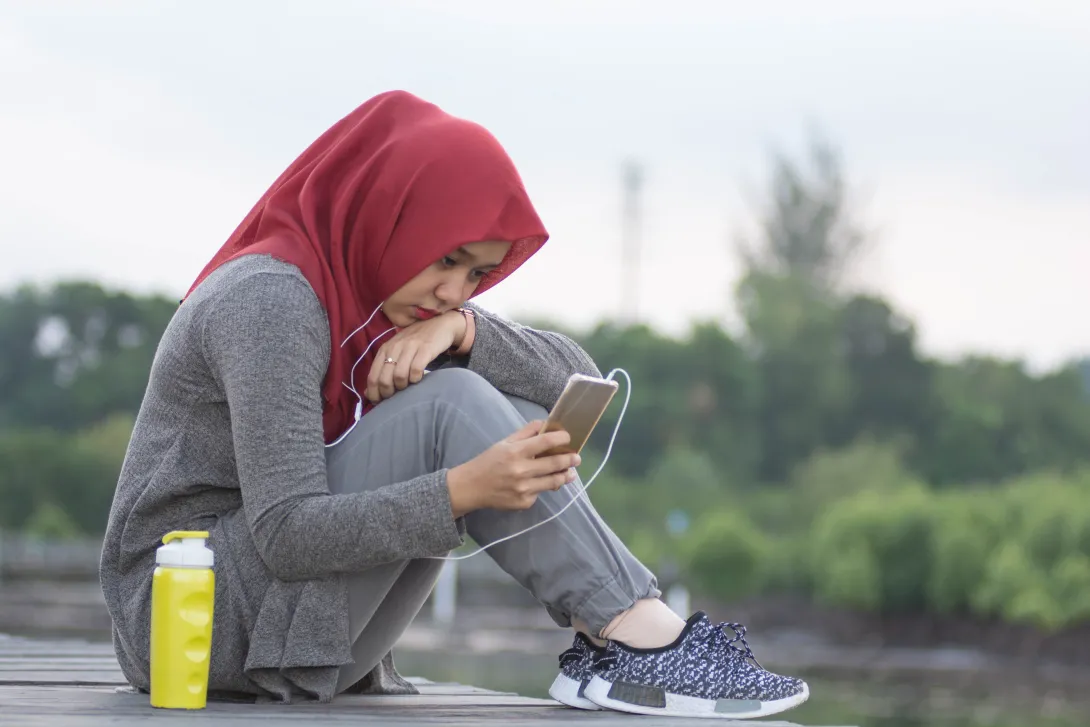 Photo of a teenager sitting with headphones in her ears and looking at her cell phone.
