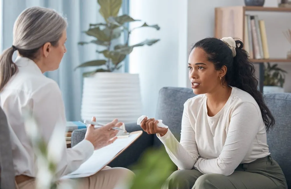 A person who appears to be upset is holding a tissue and sitting on a couch while speaking with a counselor who is taking notes on a clipboard