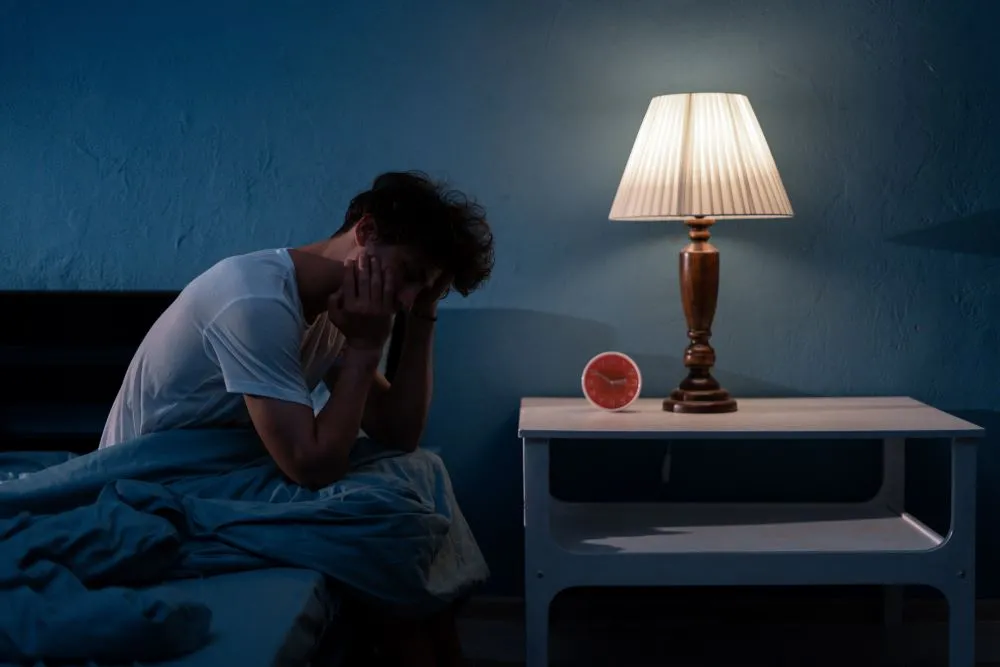 A man sitting on the edge of his bed in a dimly lit room with his hands holding his head. There is a lamp and alarm clock on the nightstand next to the bed.