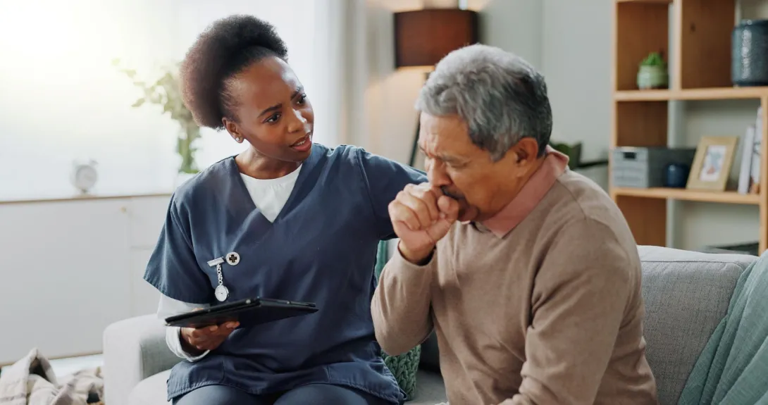 A healthcare provider offers support to a patient who is coughing into his hand while seated on a couch.